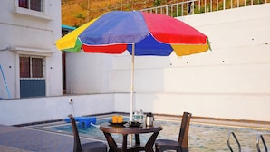 A poolside area with a table and chairs, a colorful umbrella, and a pool. - The Serenity Resort (Mahabaleshwar)