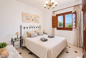 Un dormitorio ordenado con cama, mesitas de noche, lámpara, un adorno en la pared y una ventana con cortinas.