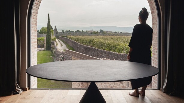 A room with a large arched window, a round table, and a person standing by the window. - Moon suite (Pescantina)