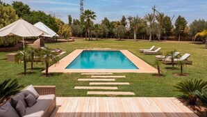 A rectangular swimming pool with a wooden deck in the foreground, surrounded by grass and palm trees. - Villa Hana B (Marrakech)