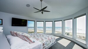 A bedroom with a bed, a ceiling fan, a TV, and large windows overlooking the beach. - Beachfront 4BR/4.5BA Luxury Getaway! (Galveston)
