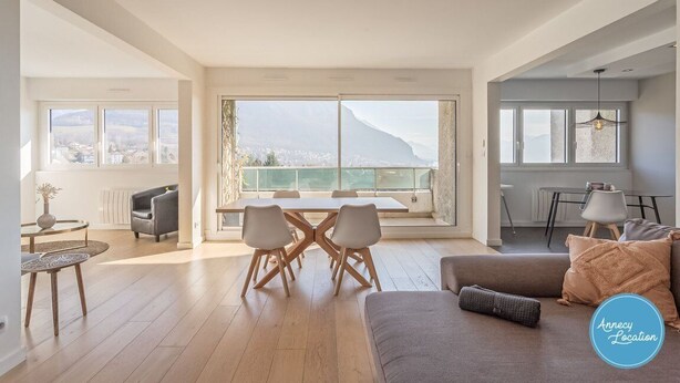 A spacious living area with a large window overlooking a cityscape and mountains. There is a dining table with chairs, a sofa, and a small side table. - Le Sainte Anne- appartement avec vue Lac. (Annecy-le-Vieux)