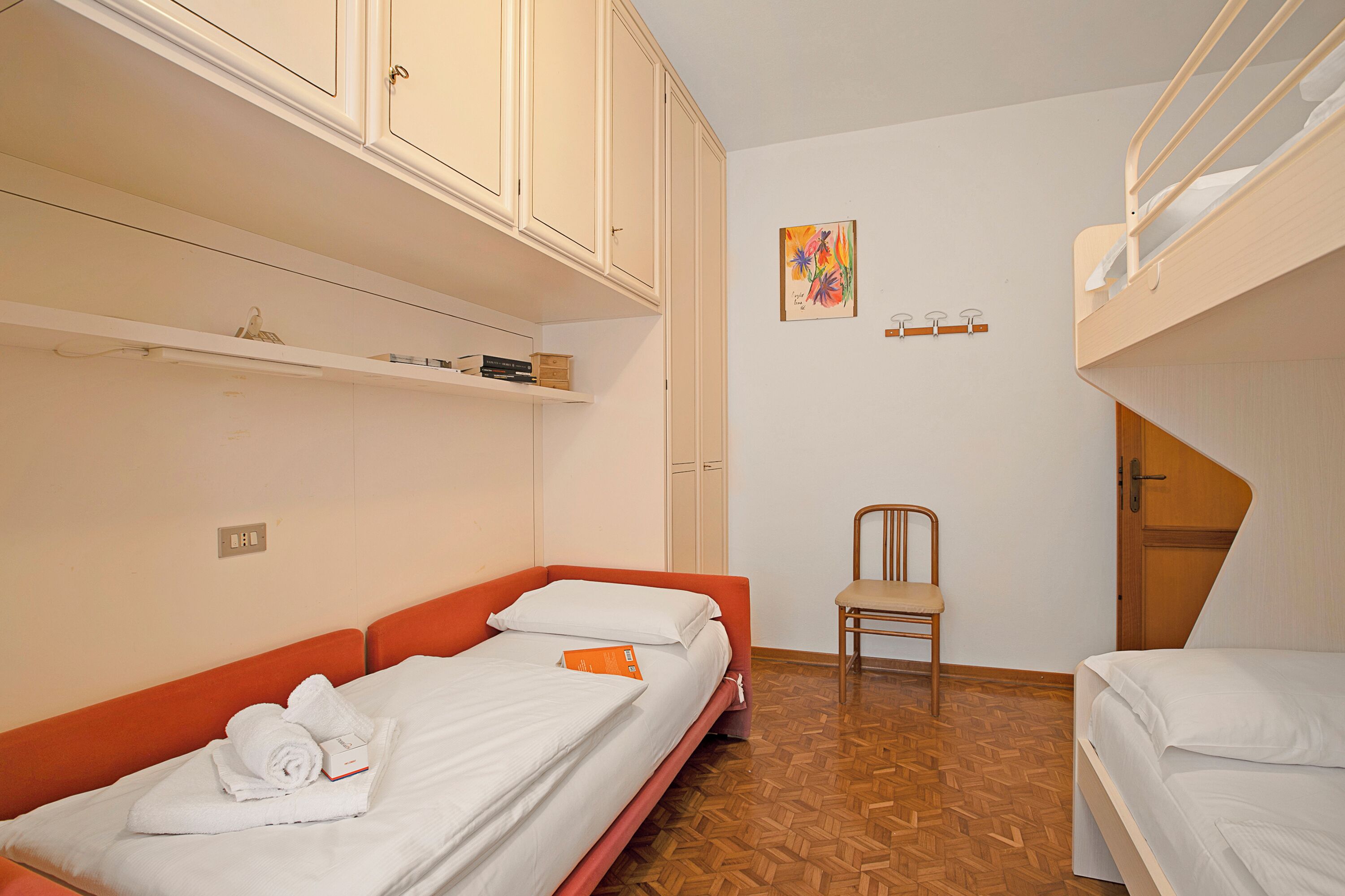 Una habitación pequeña y funcional con dos camas individuales, una silla y una litera.