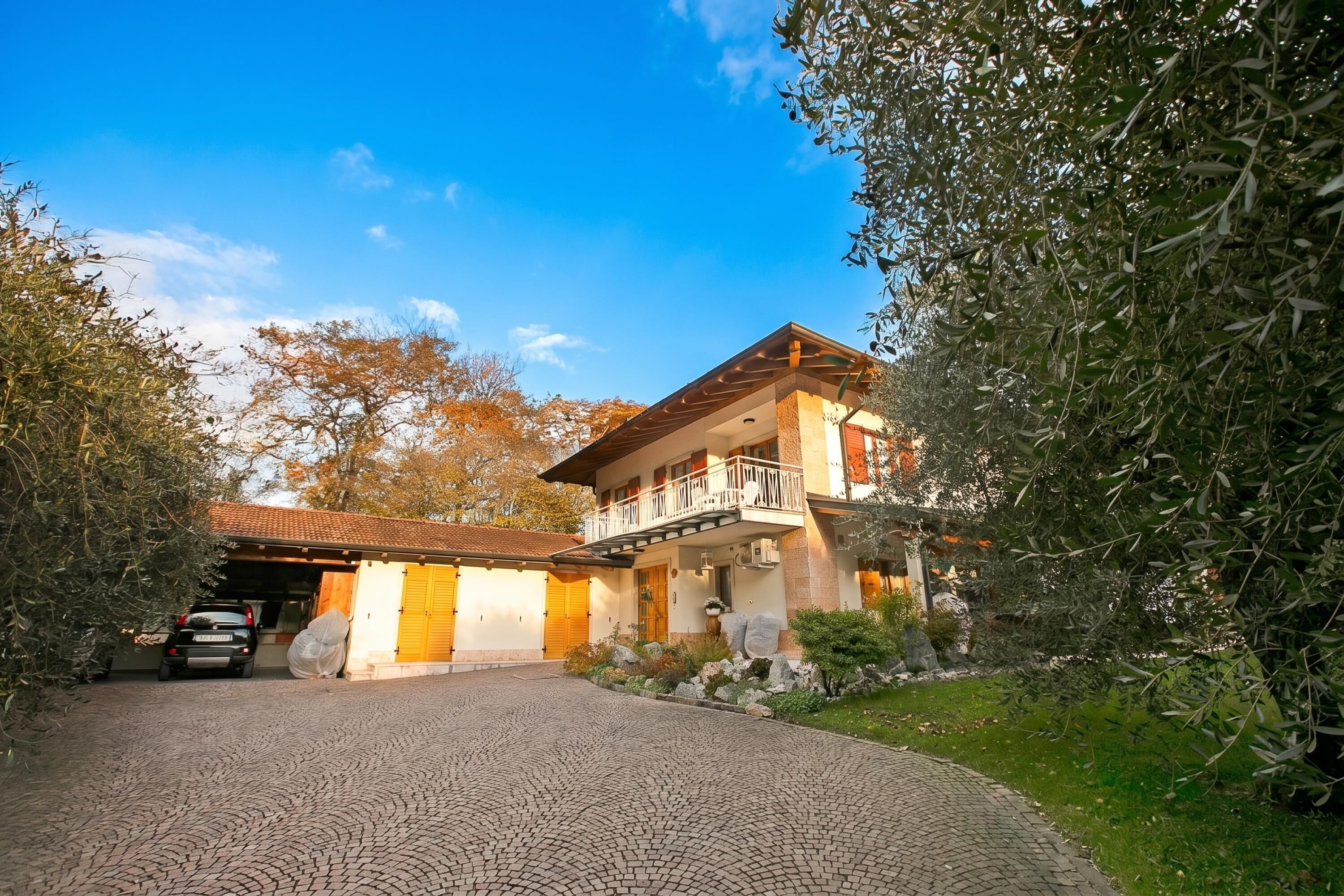 A two-story house with a balcony, a paved driveway, and surrounding greenery.