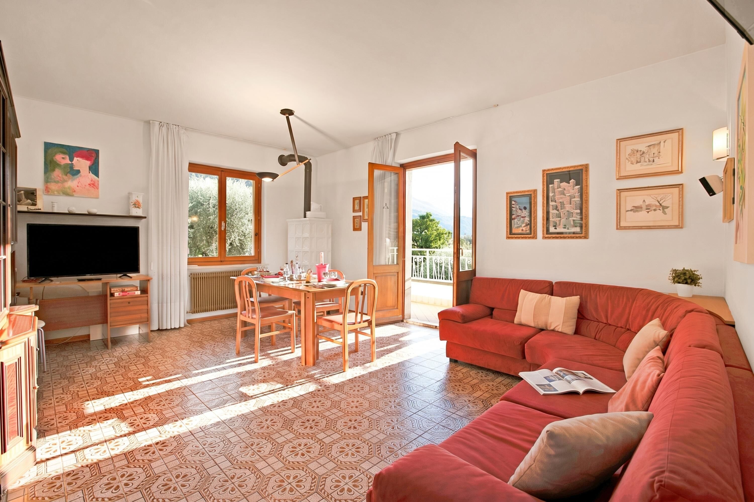 A living room with a red sofa, a TV, a dining table with chairs, and a balcony with a view.