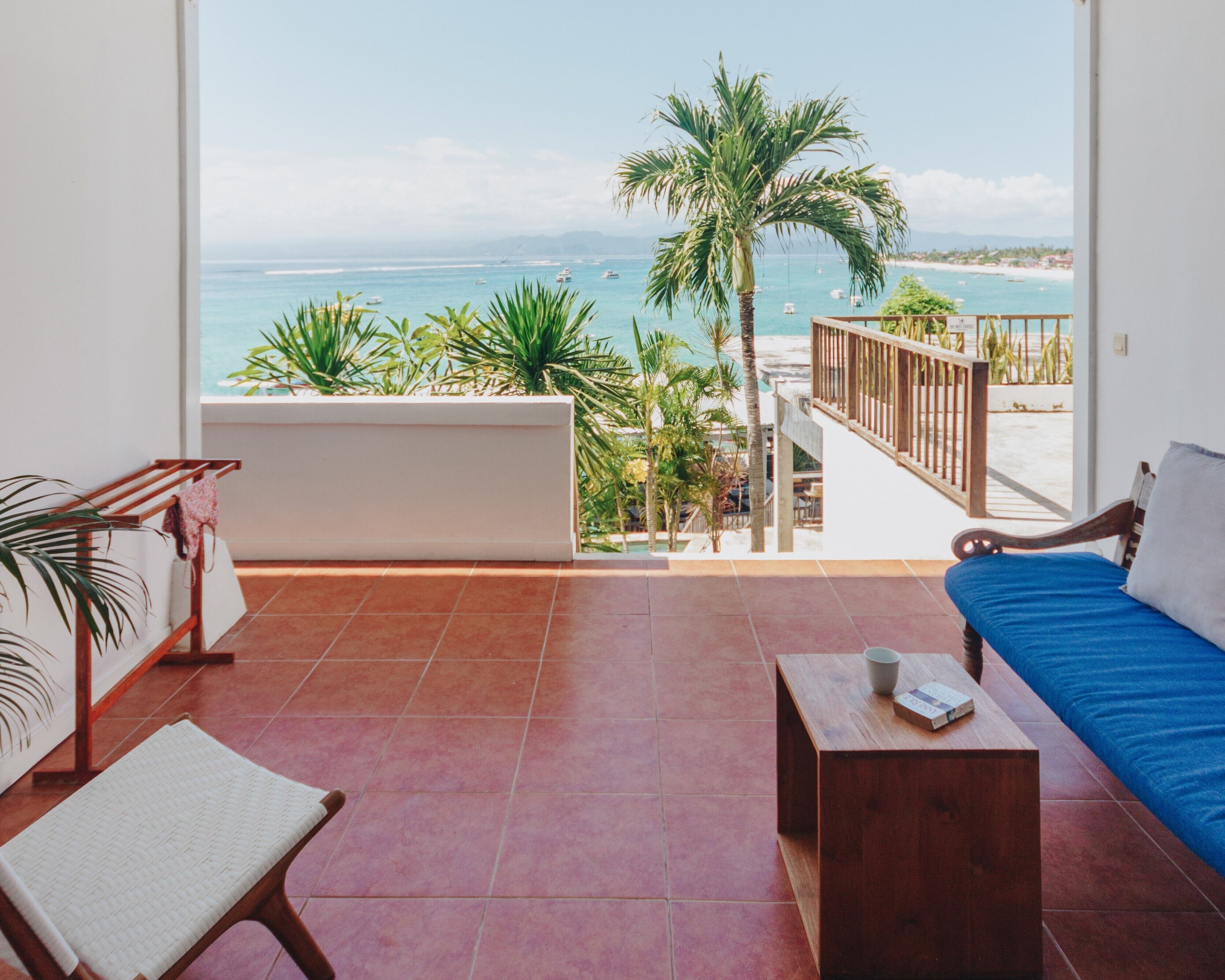 A balcony with a view of the ocean, a wooden bench, a blue sofa, and a small table with a book and a cup.