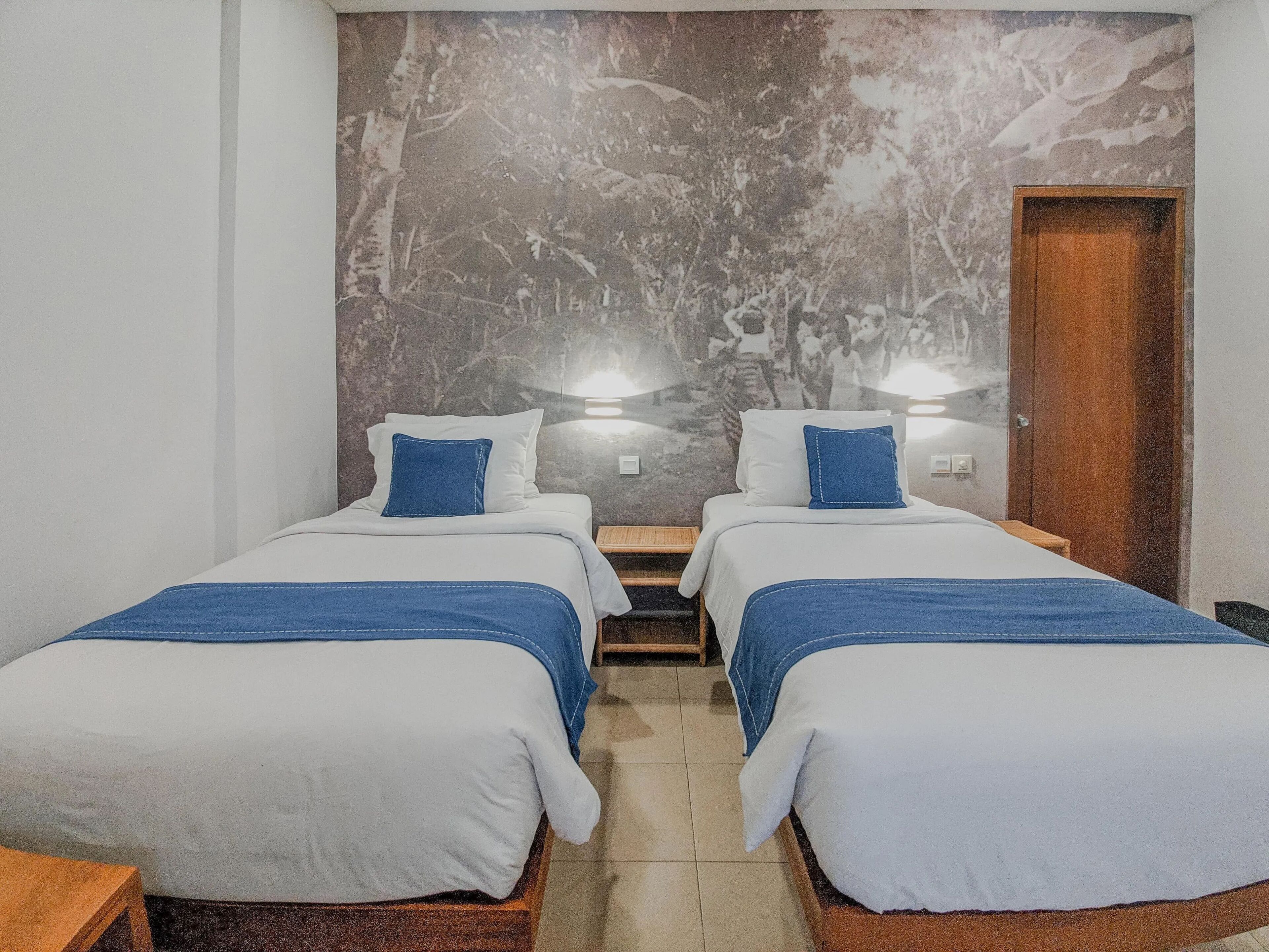 A hotel room with two beds, each with a blue blanket and white bedding. There are wall-mounted lights above each bed, and a wooden door on the right.