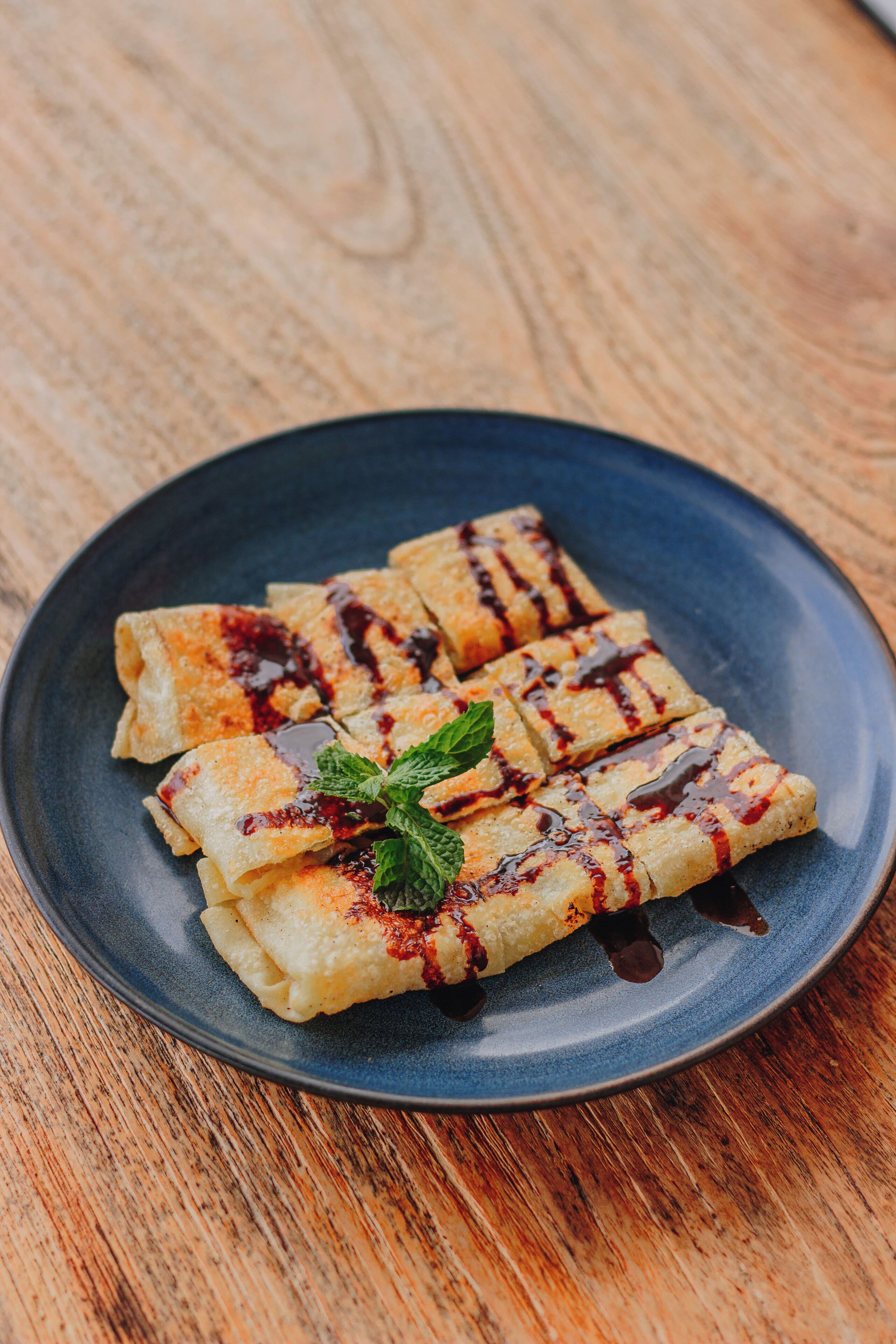 A plate of crepes with chocolate sauce and mint garnish on a wooden table.
