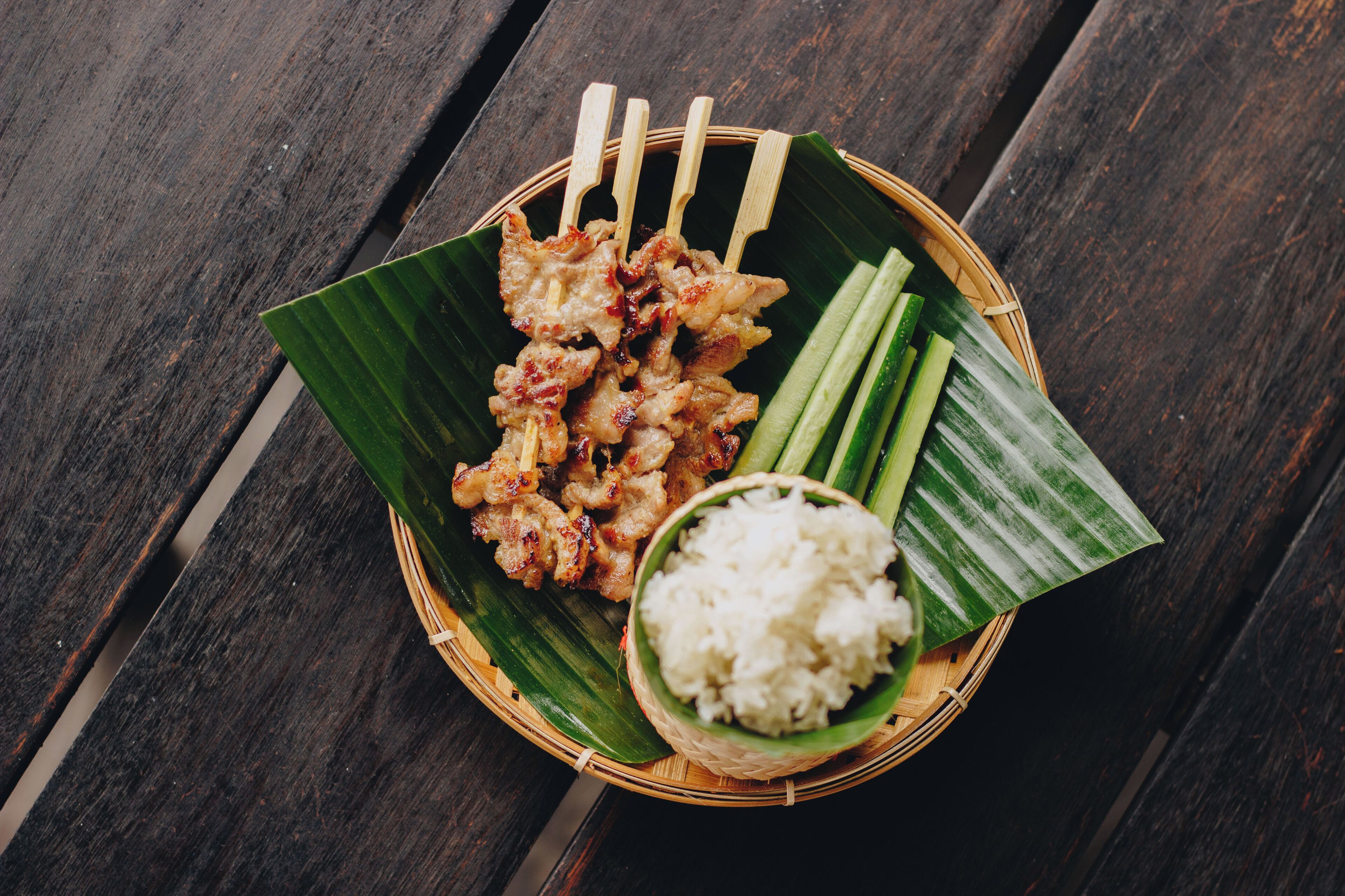 A meal of grilled skewers, rice, and cucumber slices on a wooden table.