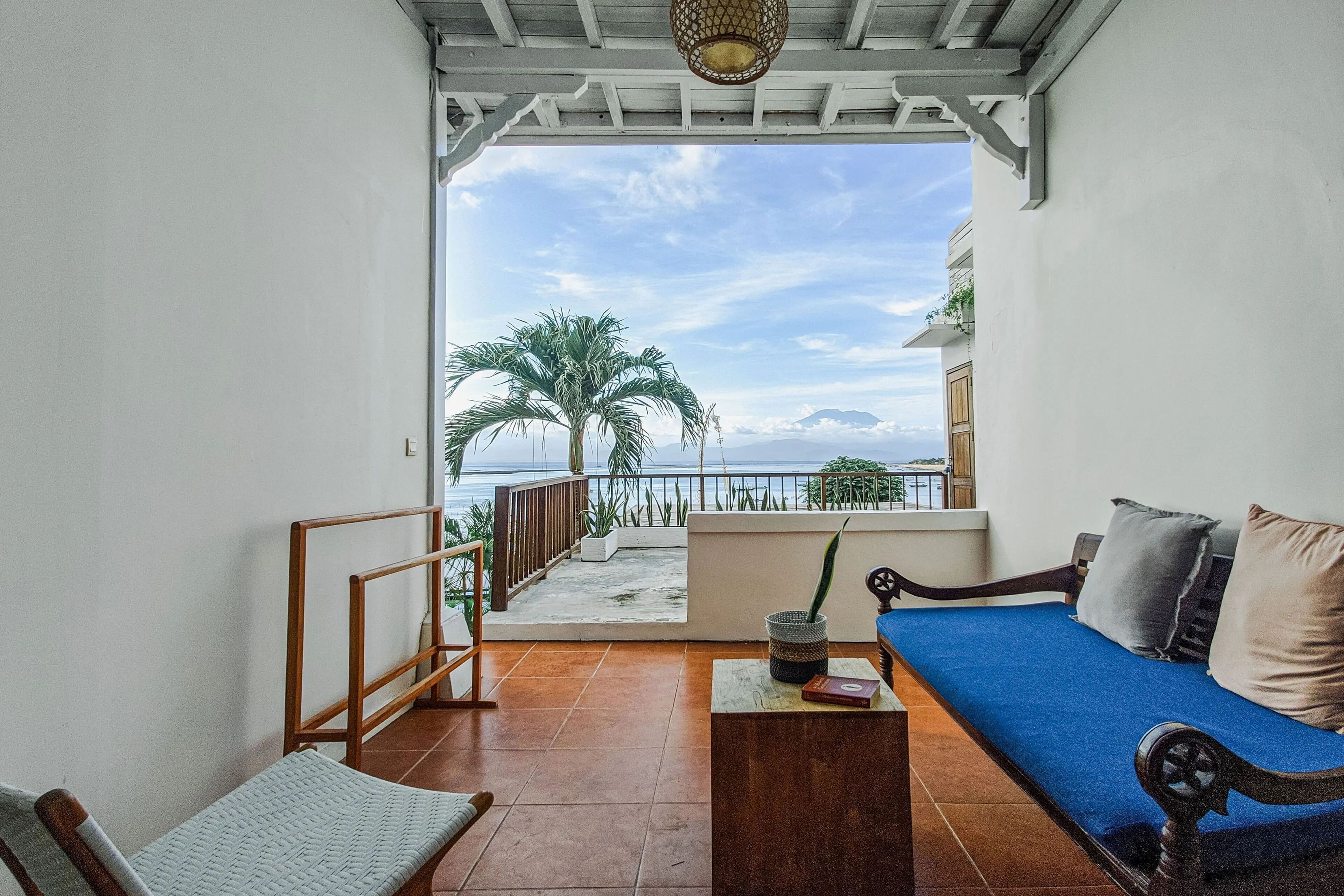 Un balcón con un sofá azul, una silla de madera y una mesita con libros. Se ve una palmera y una vista del mar a través de la barandilla.
