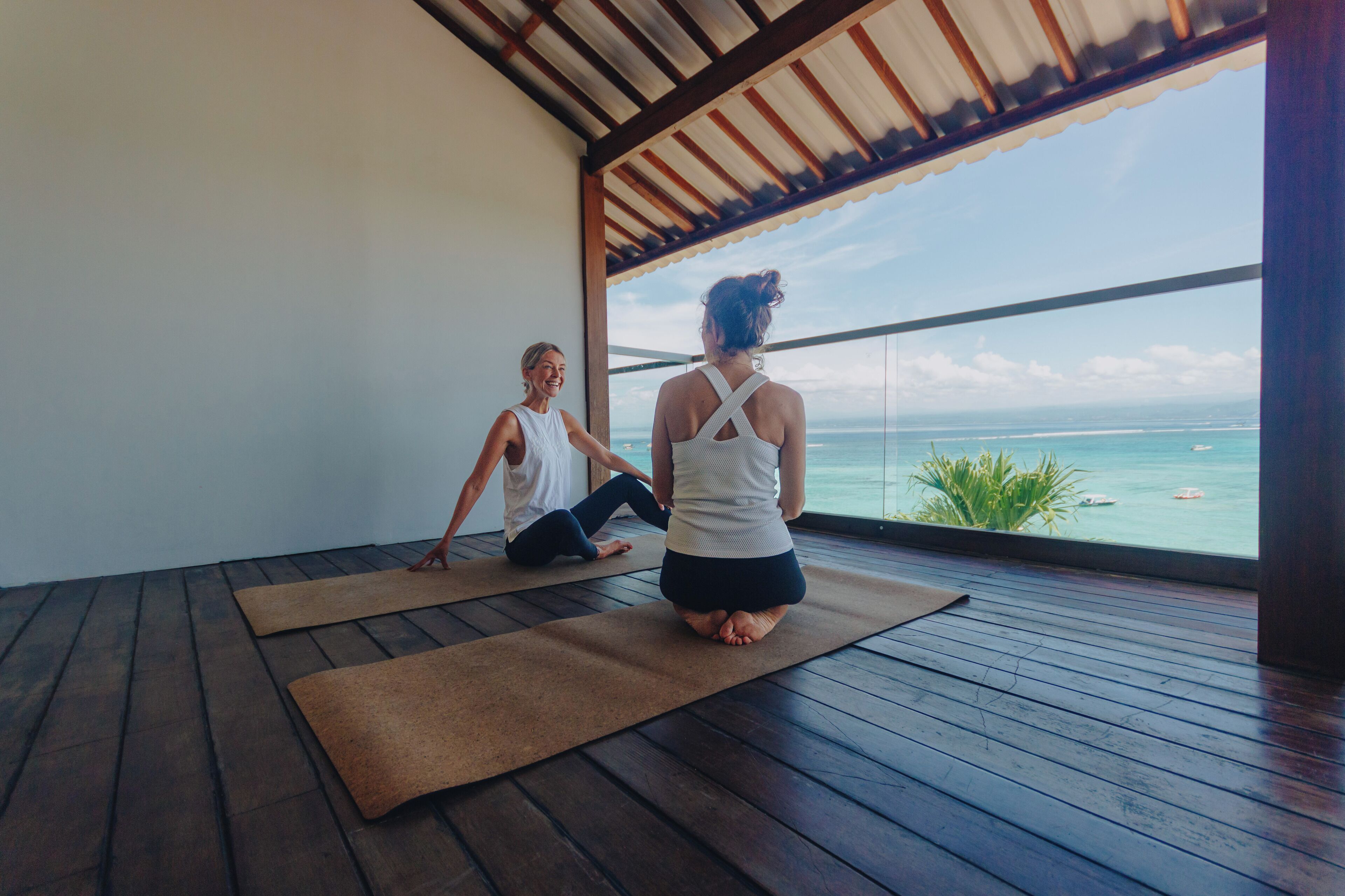 Two people doing yoga on mats in a room with a wooden floor and a large window overlooking the ocean.