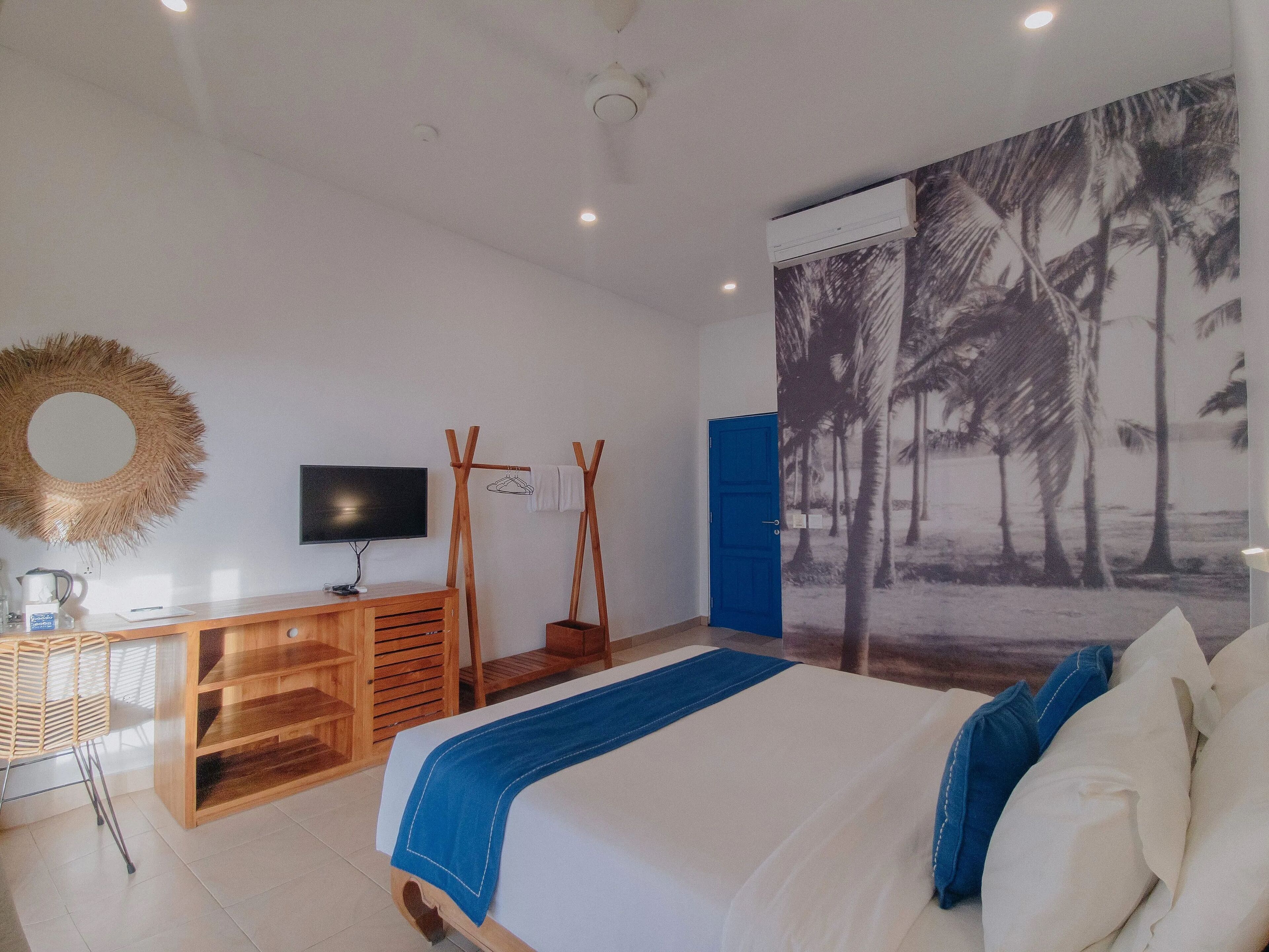 A bedroom with a bed, a TV, a wooden dresser, a mirror, and a wall with a tropical mural.