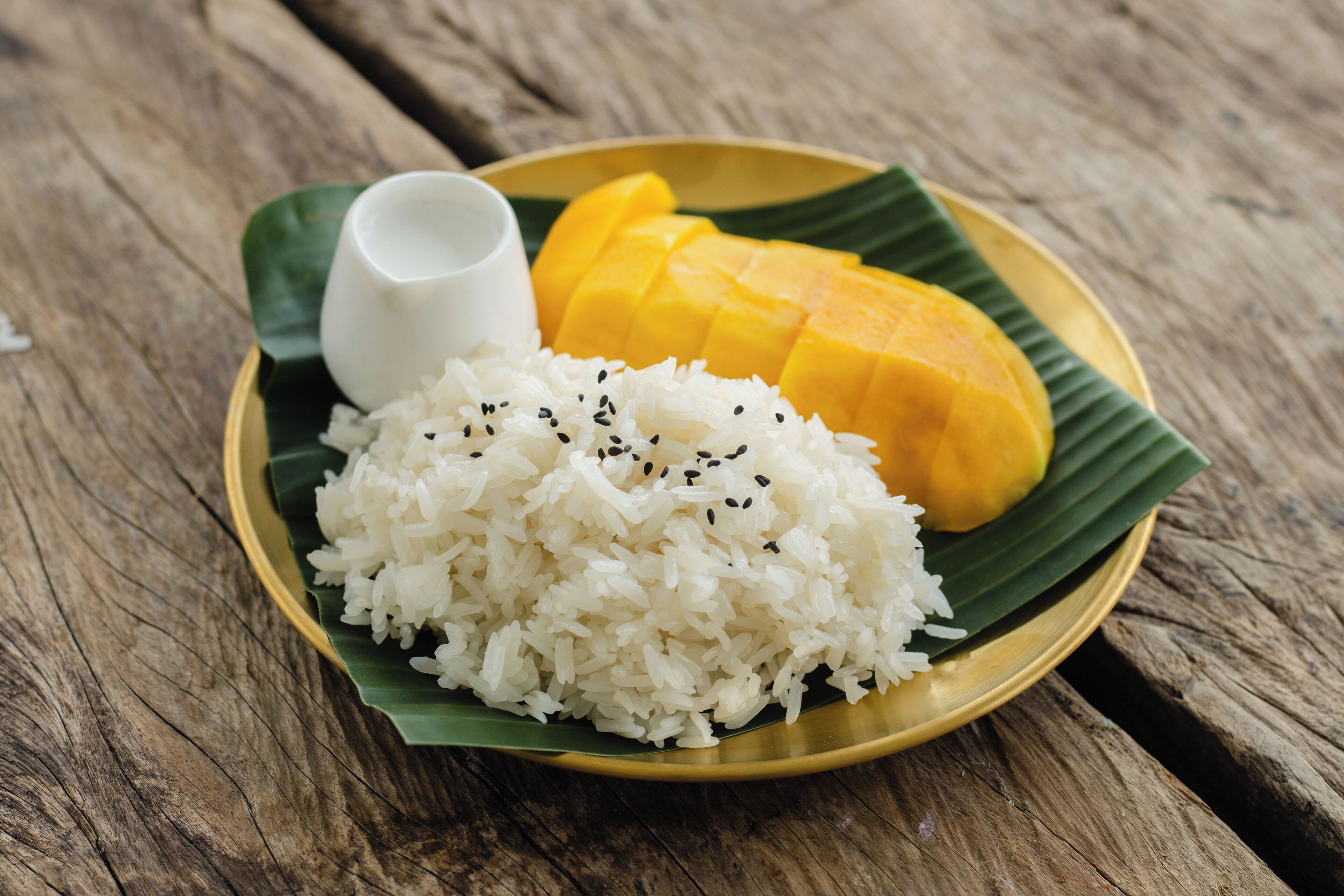 Un plato de arroz blanco con semillas de sésamo negro, un trozo de mango amarillo y una pequeña taza blanca sobre una mesa de madera.