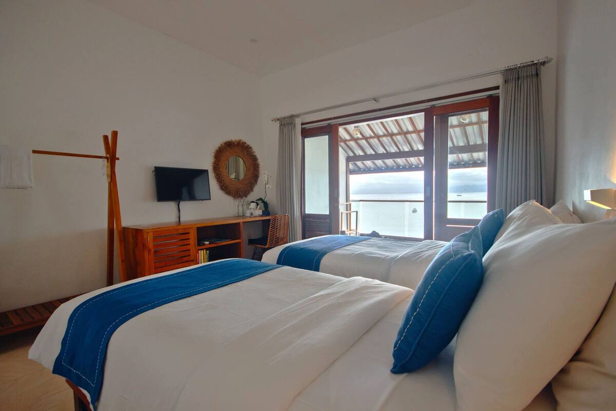 Habitació d'hotel amb dos llits, una televisió, un escriptori de fusta i vista al mar a través d'una finestra gran.