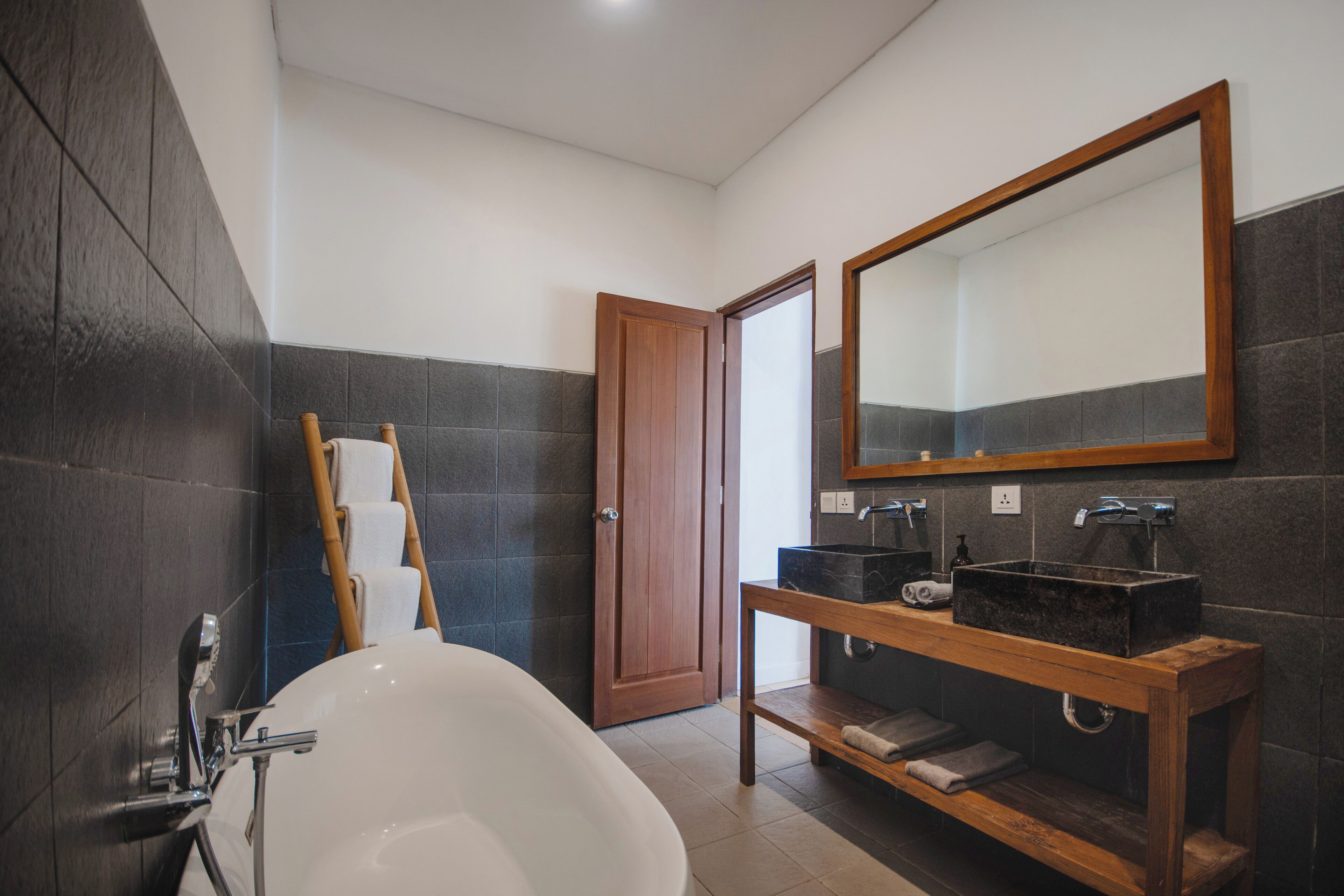 A modern bathroom with a freestanding bathtub, a wooden vanity with a large mirror, and dark tiled walls.