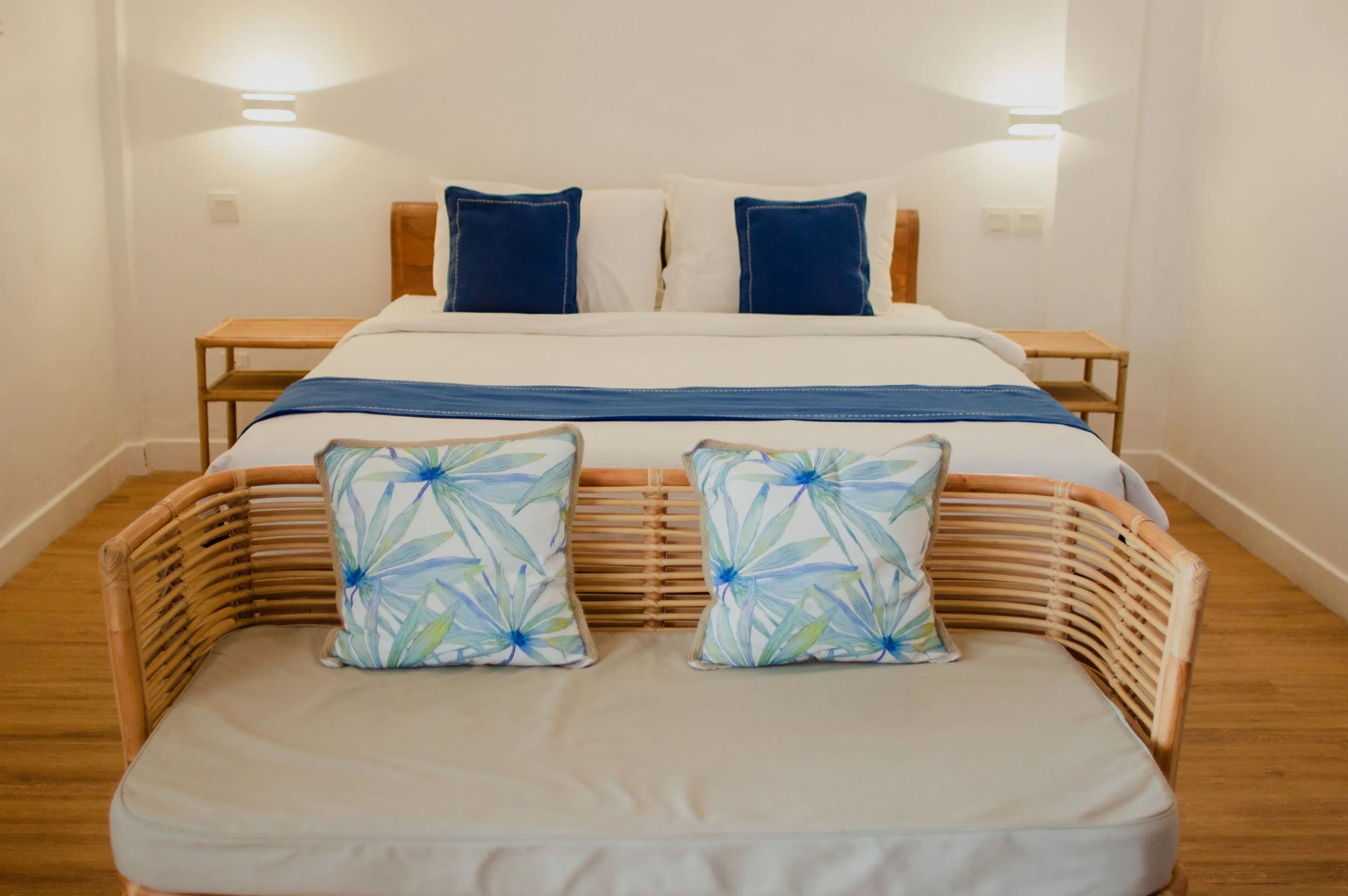 Un dormitorio ordenado con cama, dos mesitas de noche y una silla de ratán con cojines.