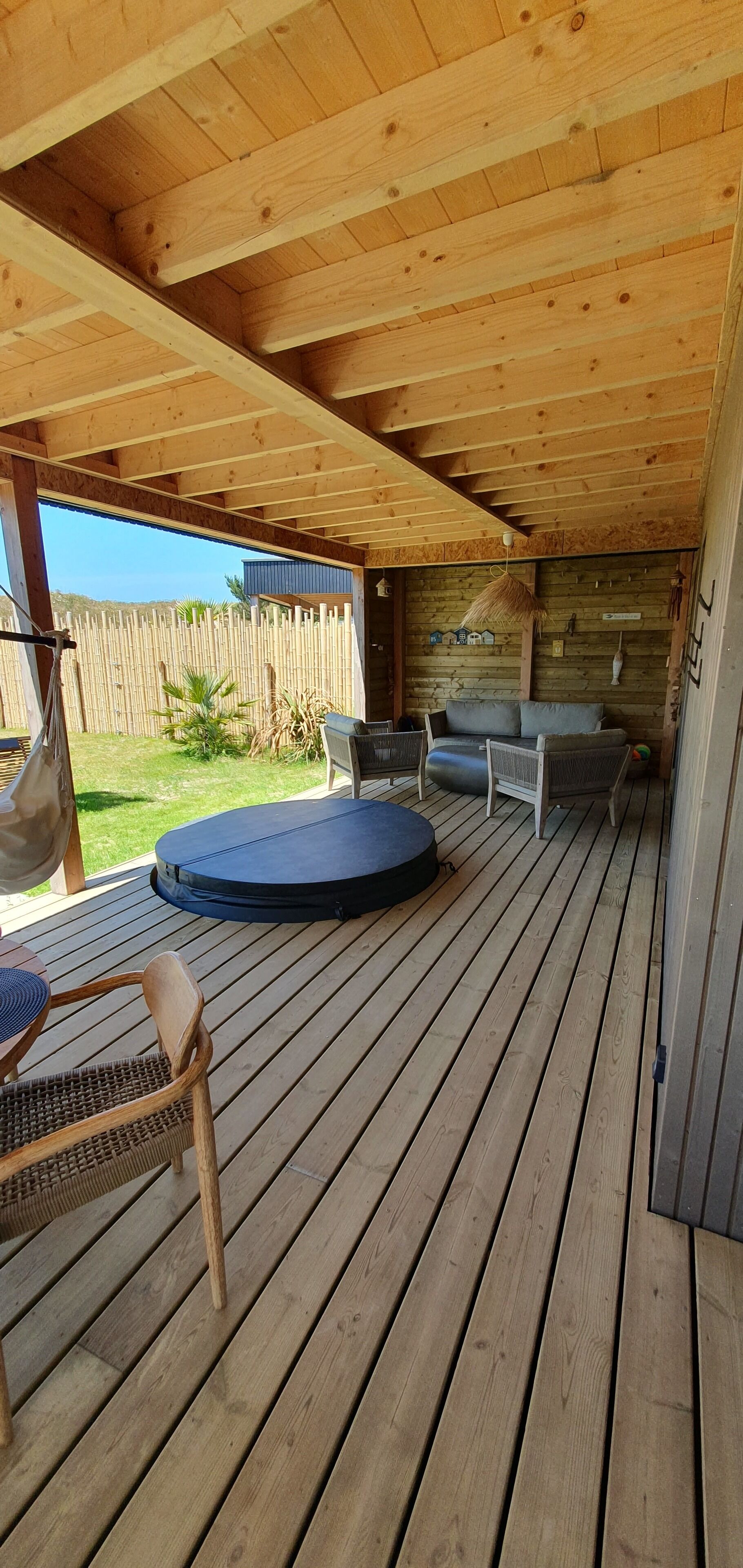 Une terrasse en bois couverte, avec un espace salon comprenant un canapé et des fauteuils, ainsi qu’un hamac.