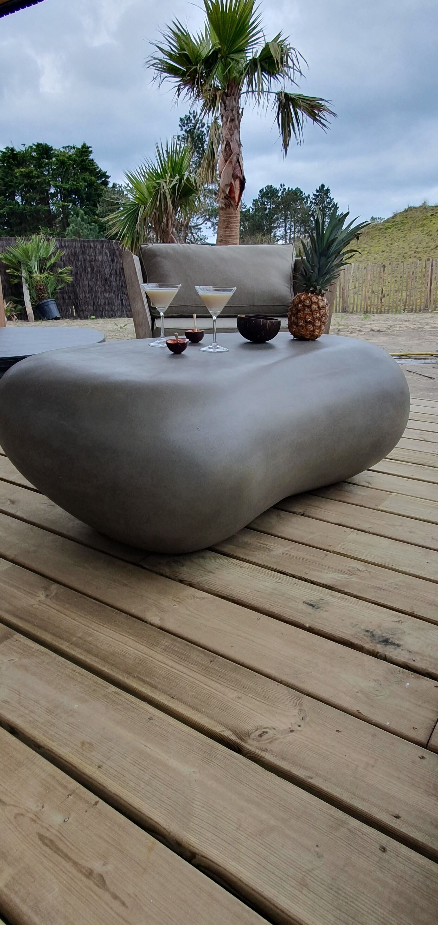 Een modern buitenzitje met een bijzondere, gebogen tafel, een ananas en twee drankjes op een houten terras.