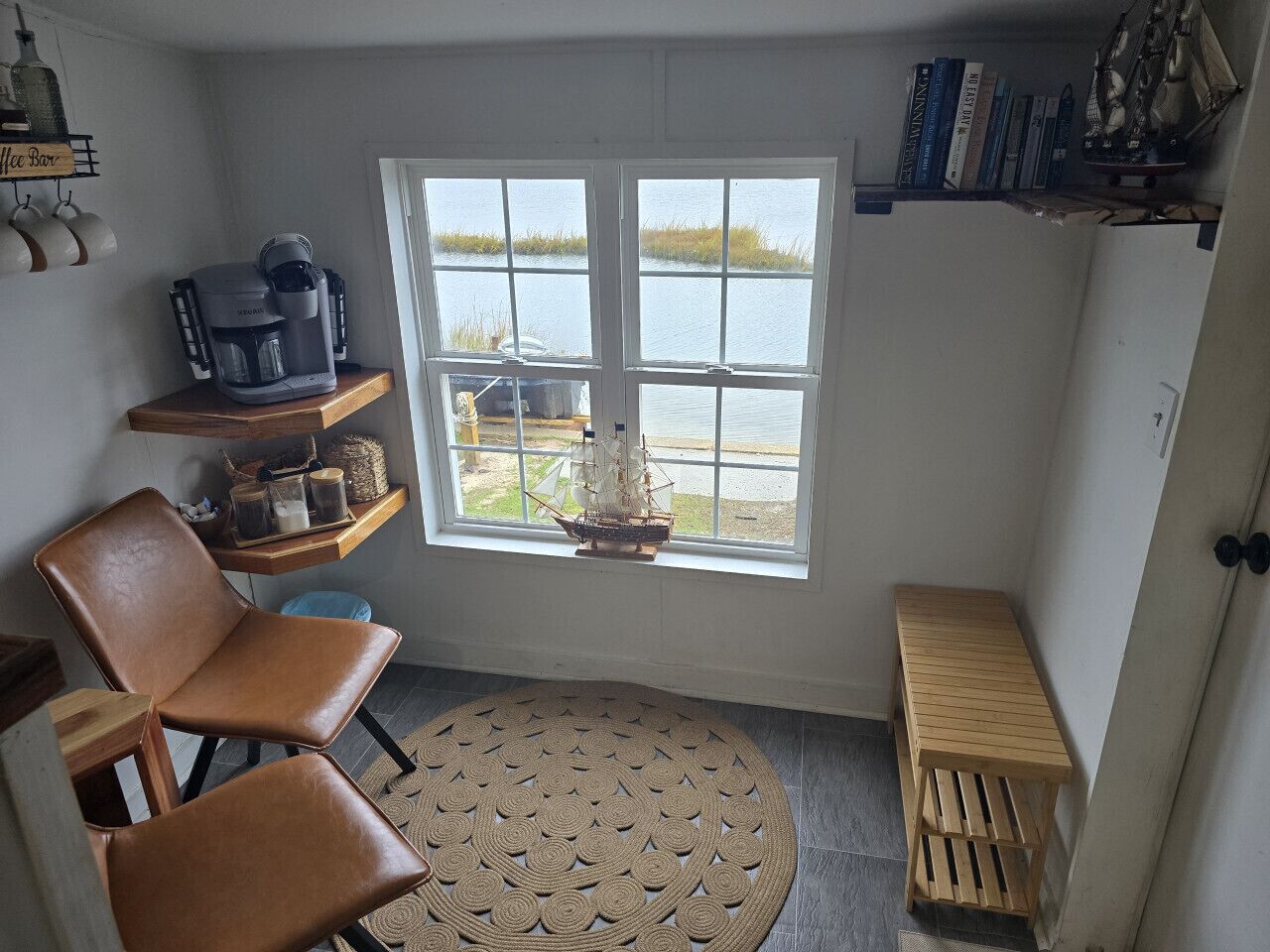 A cozy corner with a coffee maker, a chair, a small table, and a view of a body of water through a window.