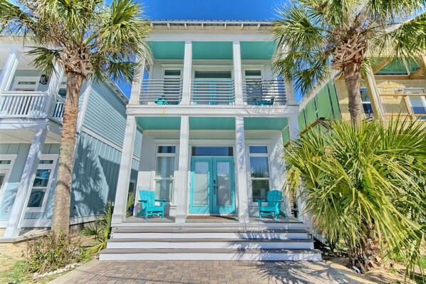 A two-story beachfront property with a balcony, turquoise chairs, and palm trees.
