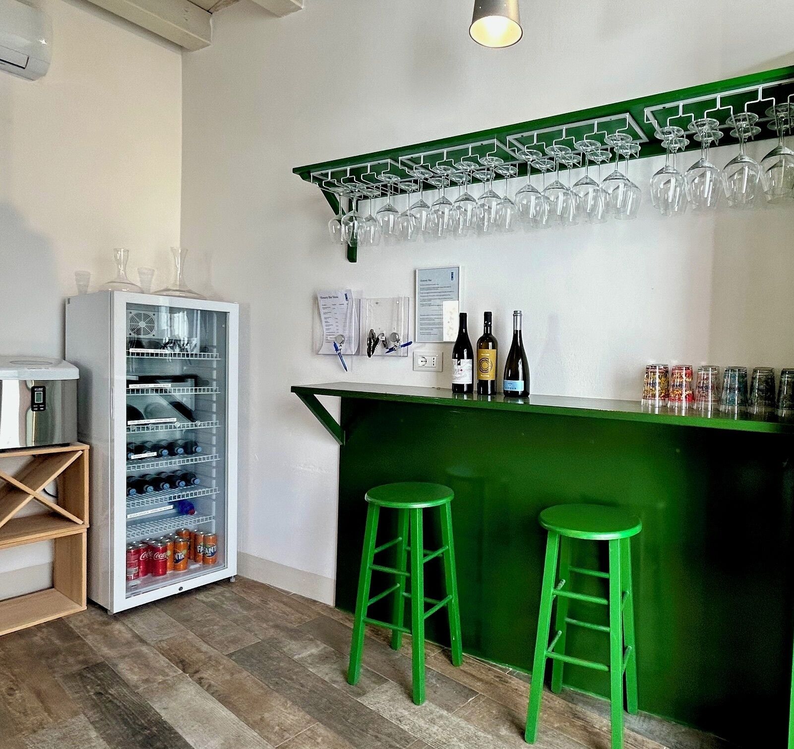A bar area with a green counter, two stools, a refrigerator stocked with beverages, and a wine rack with glasses.