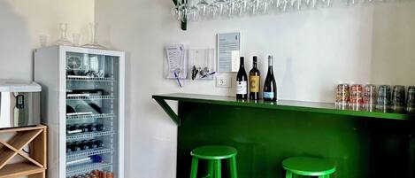 A bar area with a green counter, two stools, a refrigerator stocked with beverages, and a wine rack with glasses.