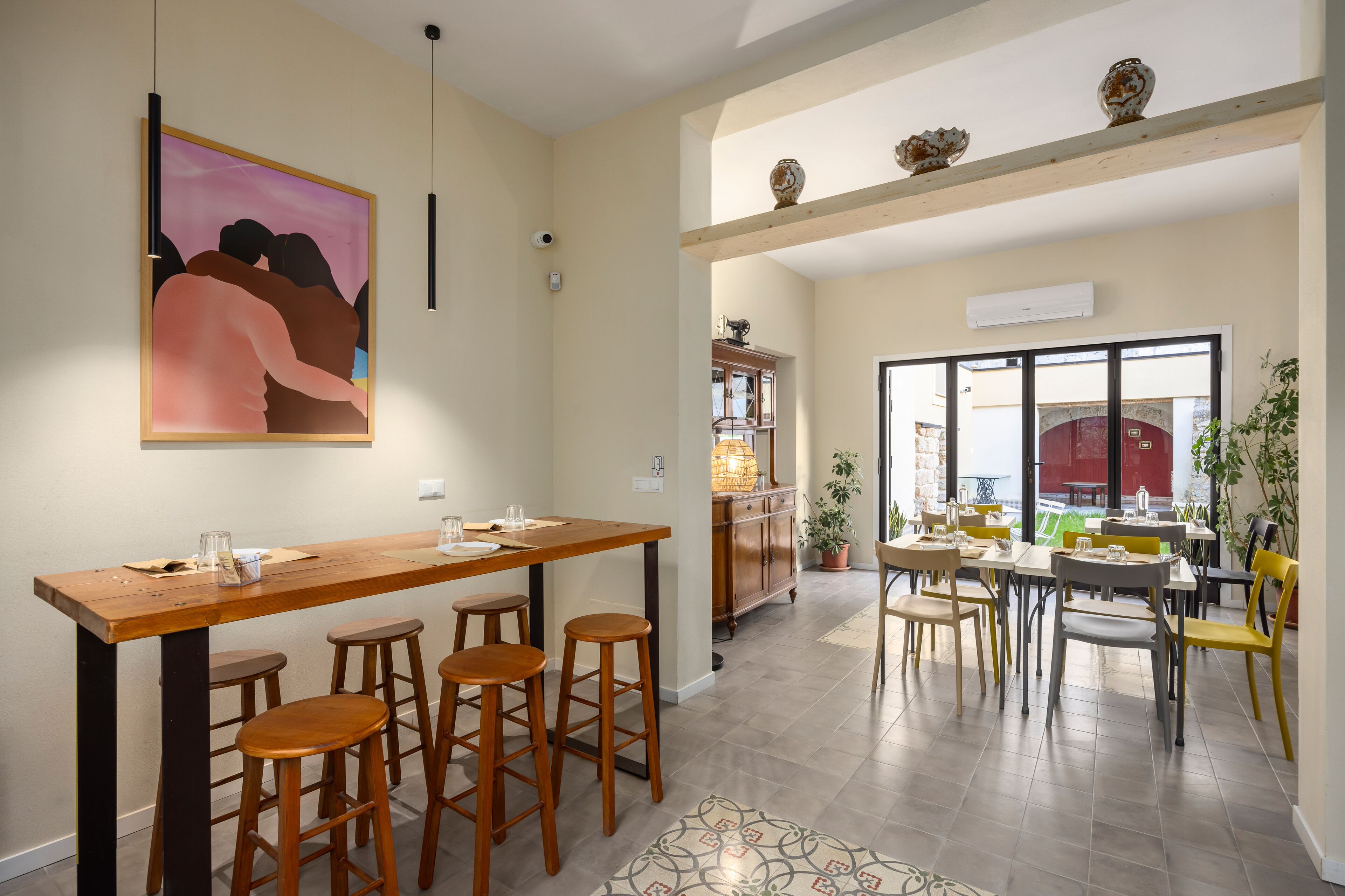 A dining area with a wooden table and bar stools, a kitchen in the background, and a large window providing a view of the outdoors.