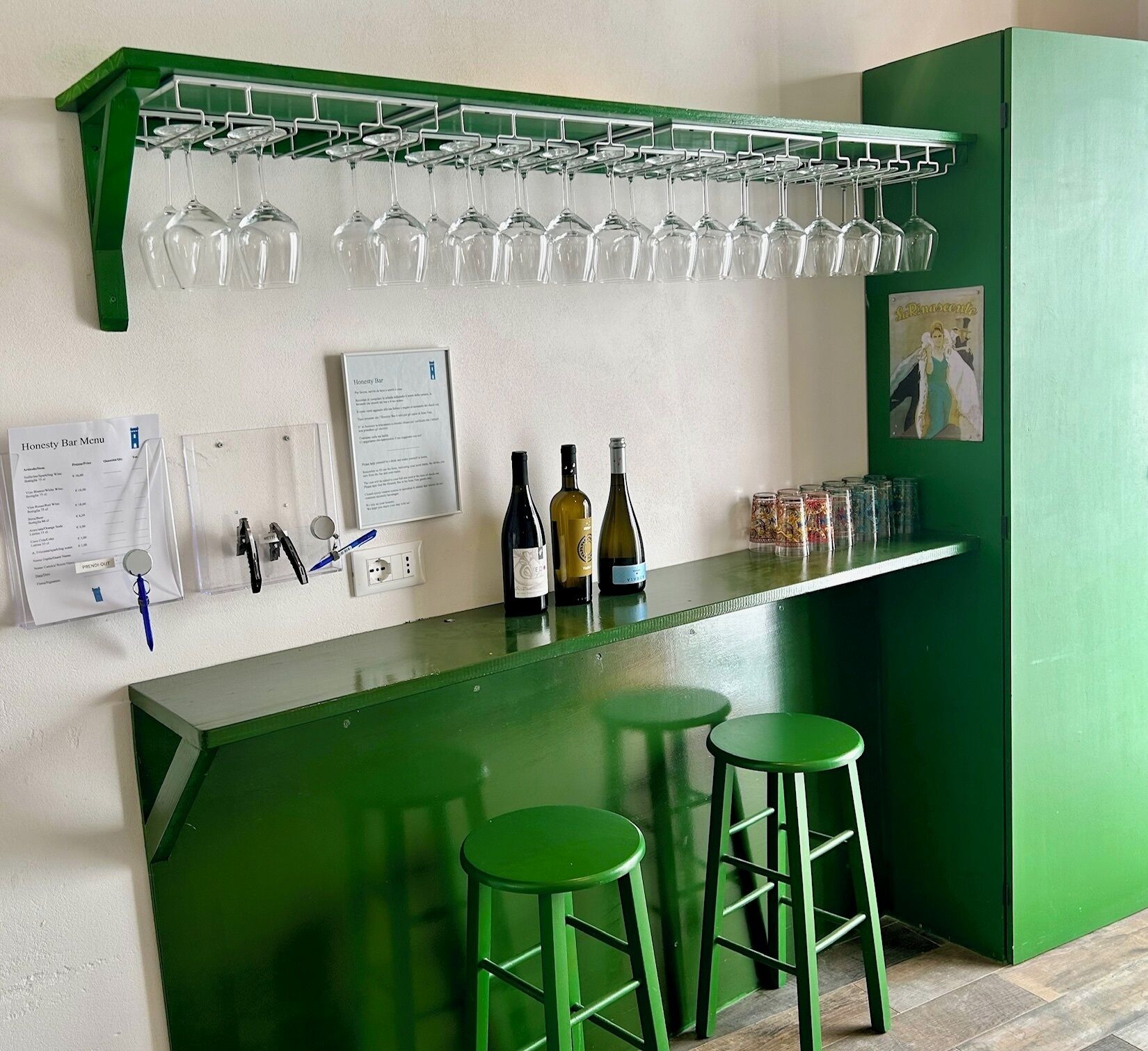 Eine Bar mit einer grünen Theke, zwei Hockern, Weinflaschen, Gläsern an einem Gestell und einer Speisekarte an der Wand.