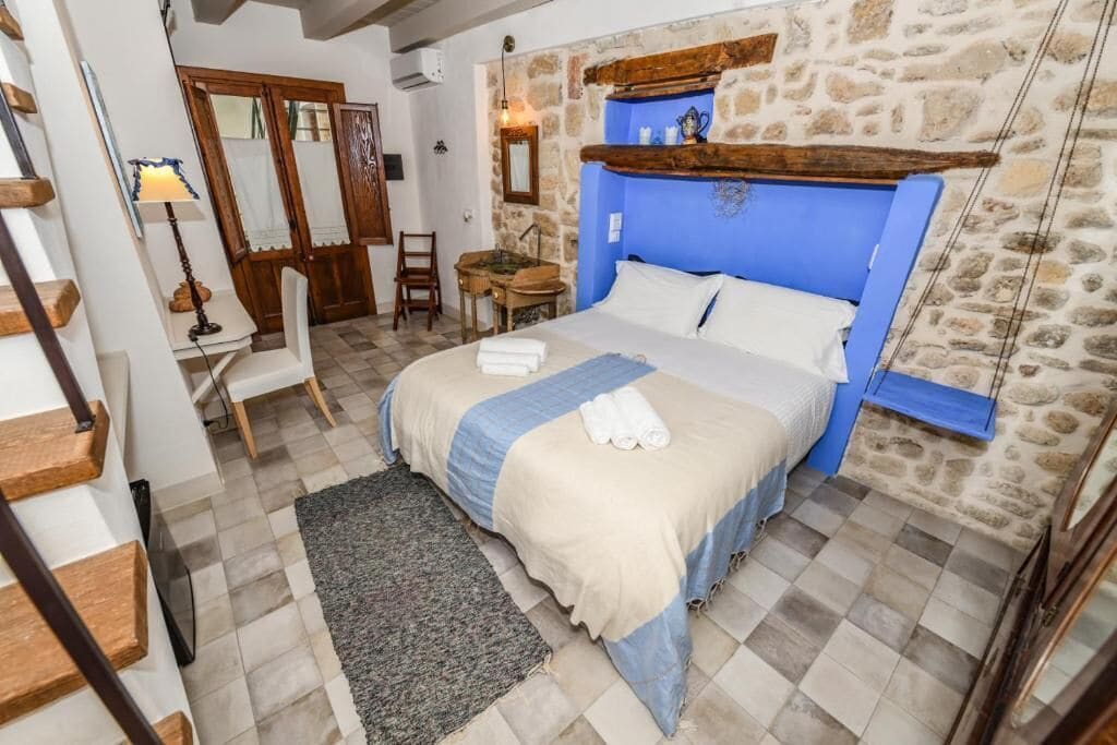 Une chambre avec un mur en pierre, une tête de lit en bois, un lit avec du linge de maison blanc, un bureau avec une lampe et une chaise.