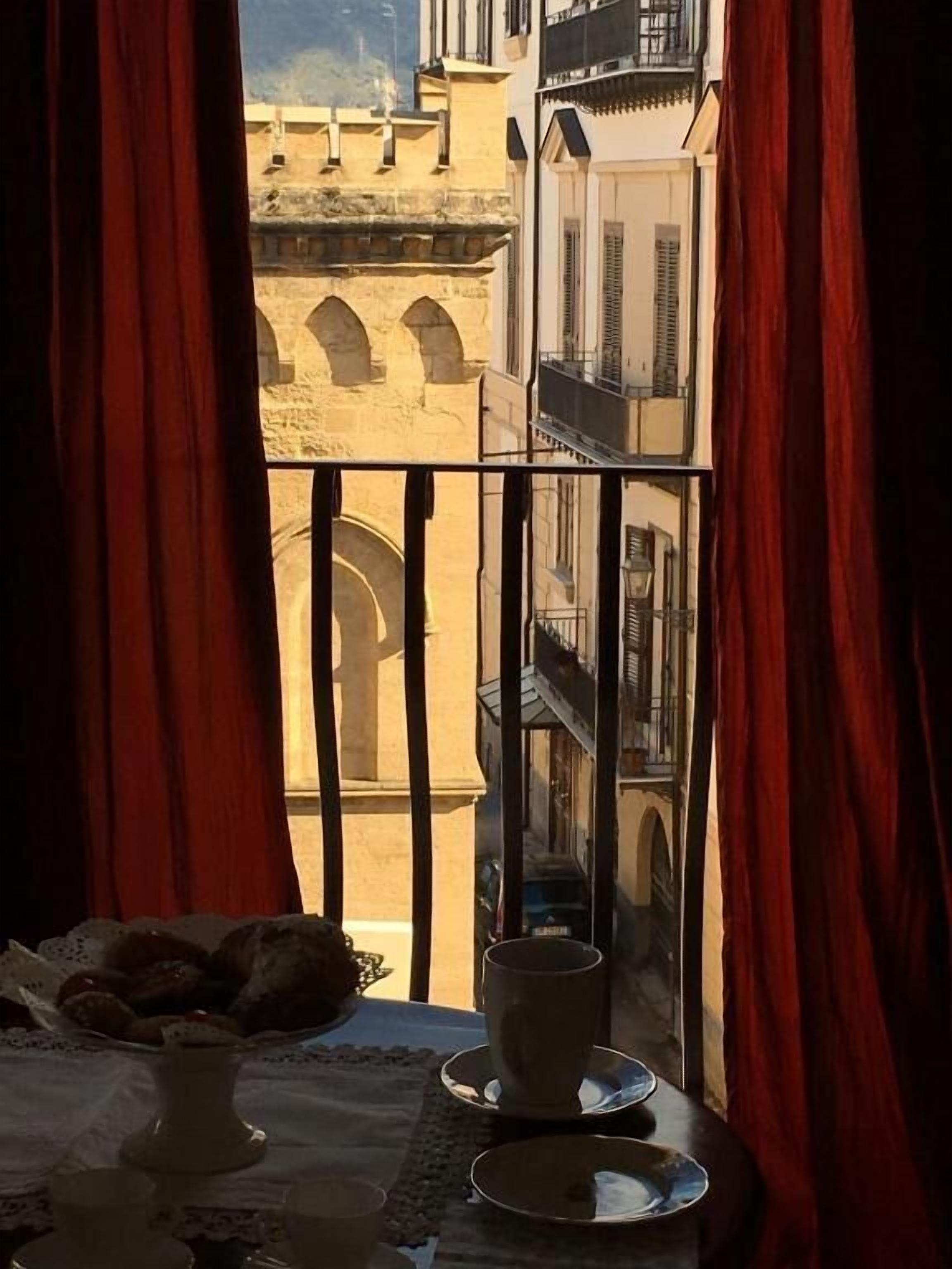 A balcony with a view of a historic building, a table with a vase and plates, and red curtains.