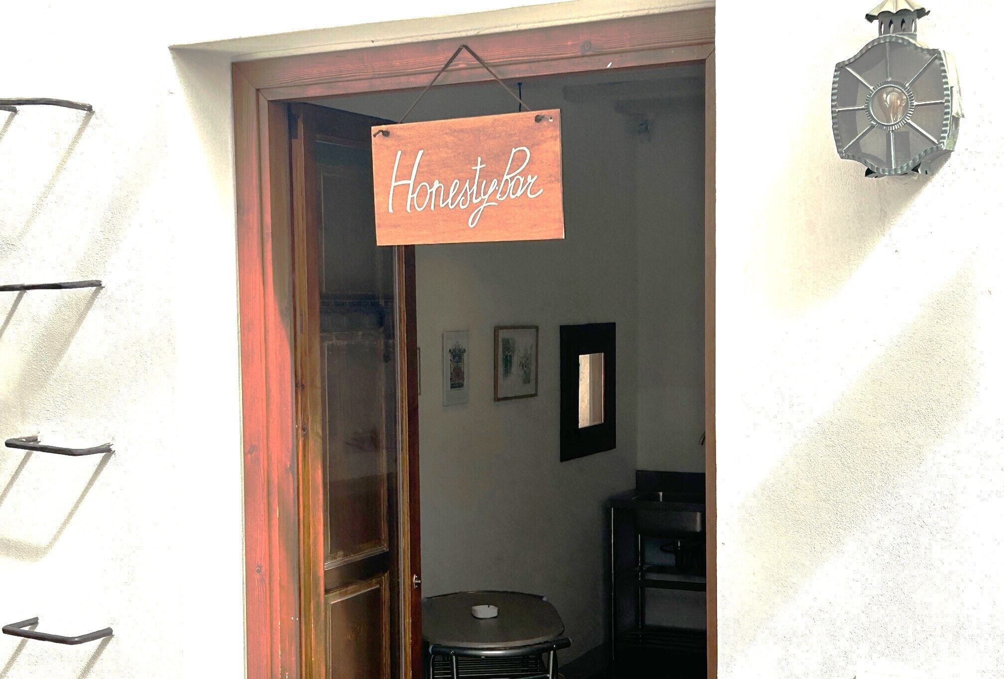 Un cartello di legno con la scritta "Honestybar" all'ingresso, un tavolino rotondo con una tazza e un lampadario a parete.