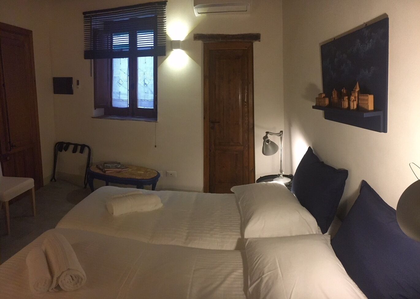 Un dormitorio con una cama grande, un escritorio, una silla, una ventana con persianas, una estantería en la pared con libros y una mesita de noche con una lámpara.