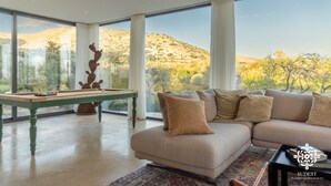 A living room with a pool table, a sofa, and a view of a hilly landscape through large windows. - Villa Dama - Luxury Escape (Avola)