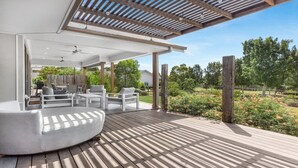 Ein überdachter Außenbereich mit Holzterrasse, weißen Möbeln und Blick auf einen Garten und Bäume.