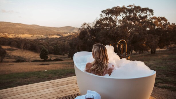 A person relaxing in a large bathtub filled with bubbles, overlooking a scenic outdoor landscape with hills and trees. - Tiny Ikigai (Whittlesea)