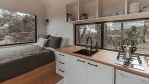 A compact kitchen with a wooden countertop, a sink, and a stove. There is a bed with a view of trees through the window. - Tiny Ikigai (Whittlesea)