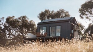 A modern house with large windows, a deck, and a solar panel on the roof, surrounded by trees and tall grass. - Tiny Ikigai (Whittlesea)