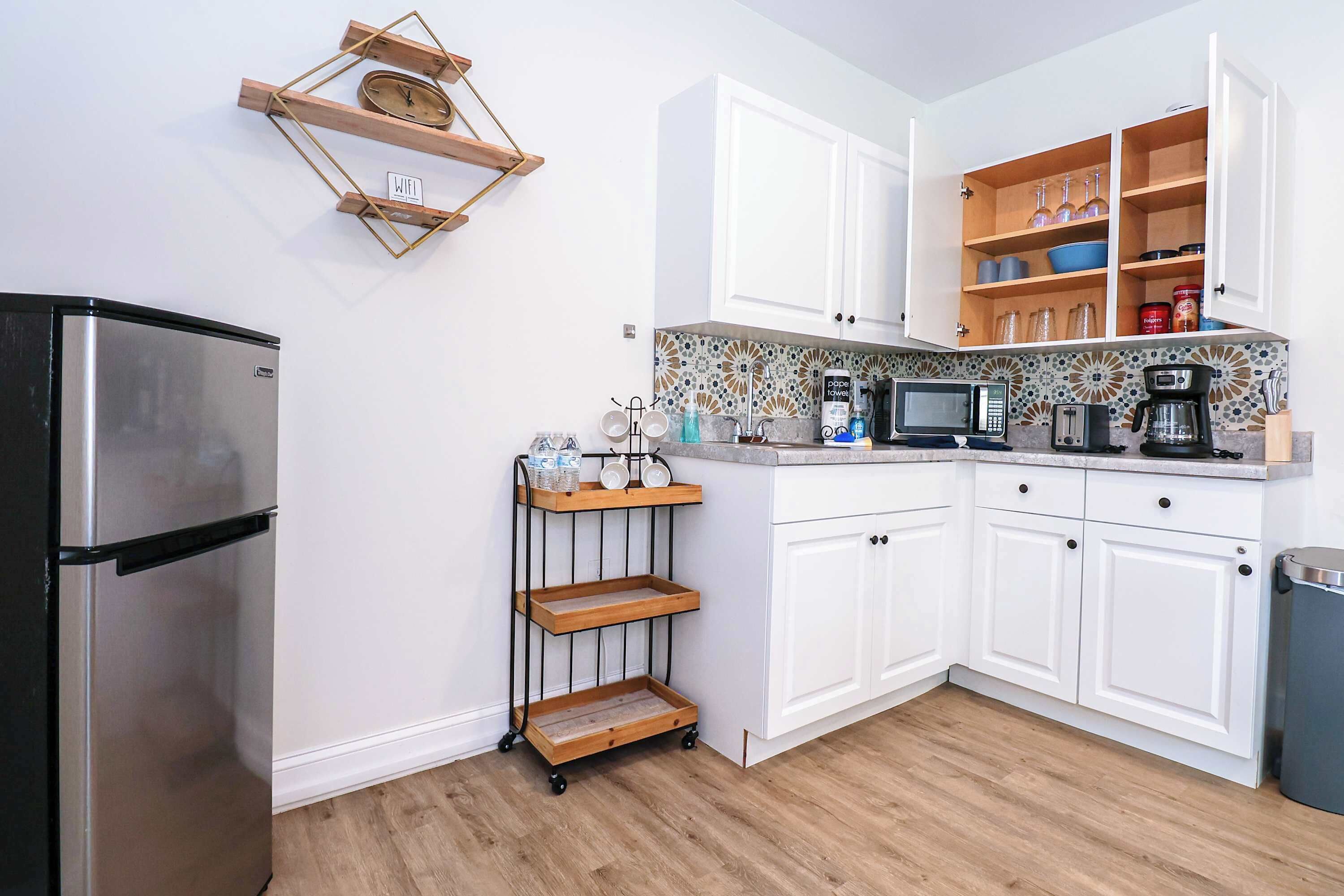 A kitchen with white cabinets, a stainless steel refrigerator, a microwave, a coffee maker, and a wooden shelf unit.