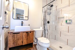 A bathroom with a wooden vanity, a white toilet, a shower with a patterned curtain, and a mirror above the sink.