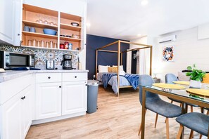Habitación de hotel compacta con kitchenette, cama con dosel, comedor con mesa y sillas, y un pequeño balcón.