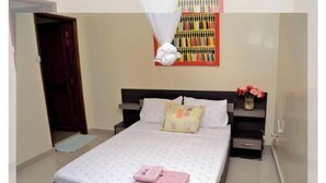 A neatly made bed with pillows and a folded towel, a bedside table with a vase of flowers, and a wall with a colorful artwork. - Chez Ouly (Dakar)