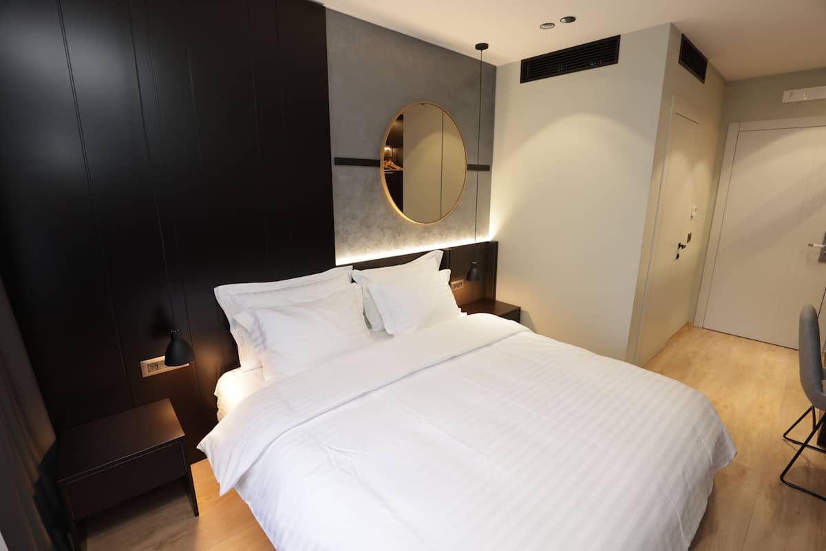 A modern hotel room with a large bed, a round mirror above the headboard, and a small bedside table.
