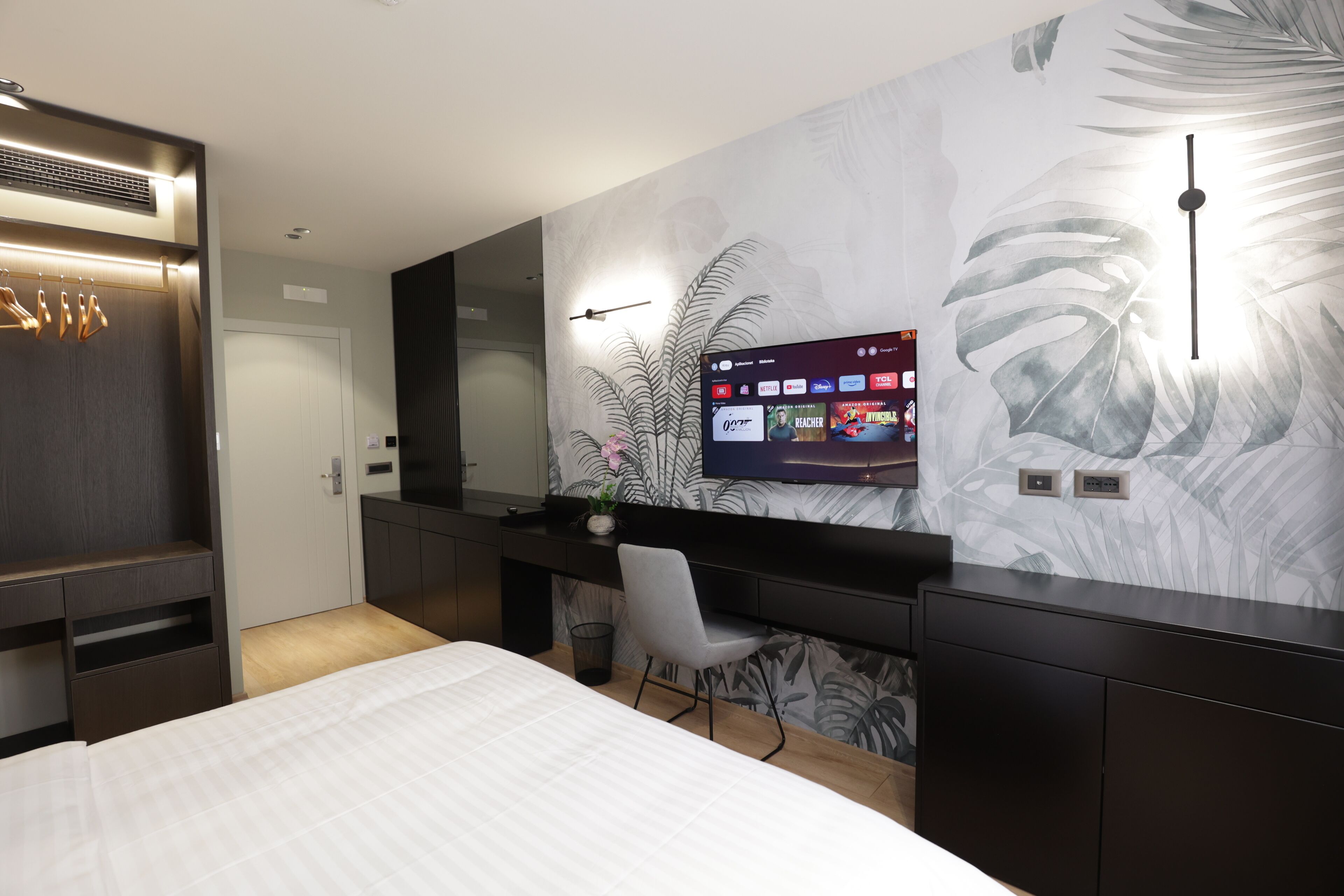 A modern hotel room with a bed, a desk with a computer, a large mirror, and a wall with a leaf pattern.