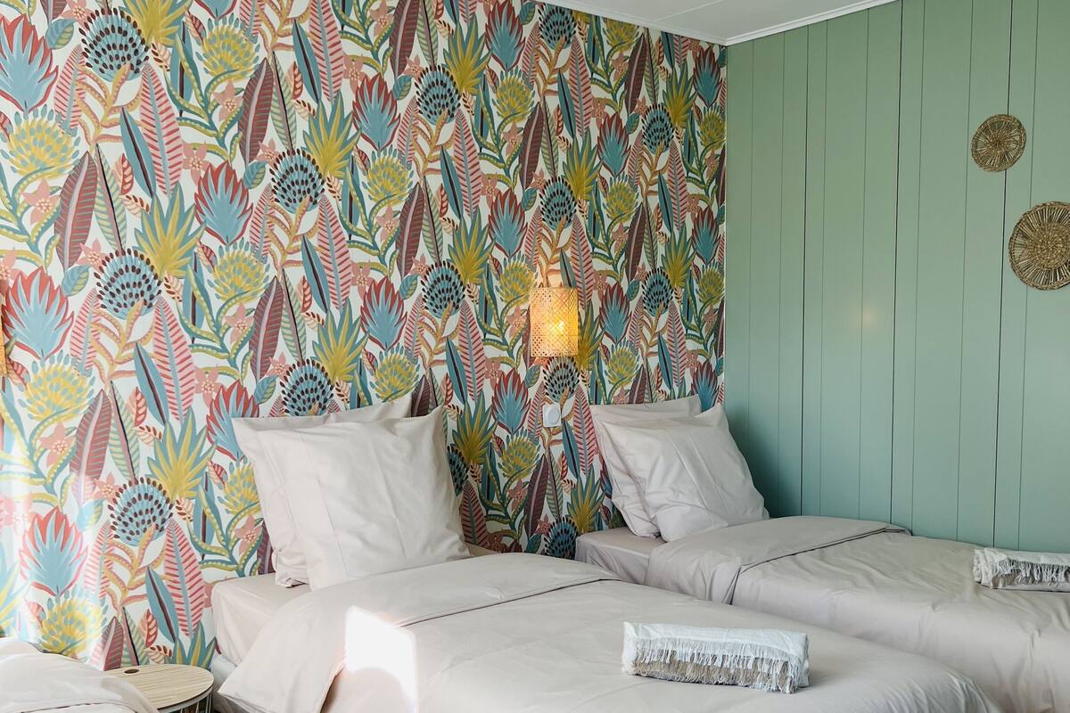 Une chambre avec trois lits, un mur aux couleurs vives orné d’un motif de feuilles tropicales, une petite table ronde et un luminaire fixé au mur.