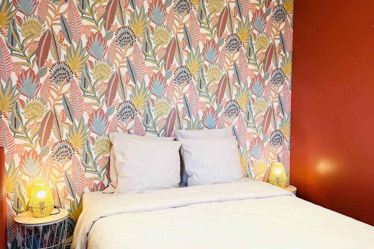 Une chambre à coucher avec un papier peint à motifs, un lit recouvert d’une literie blanche, deux lampes de chevet et une petite table ronde sur laquelle se trouve une lampe.