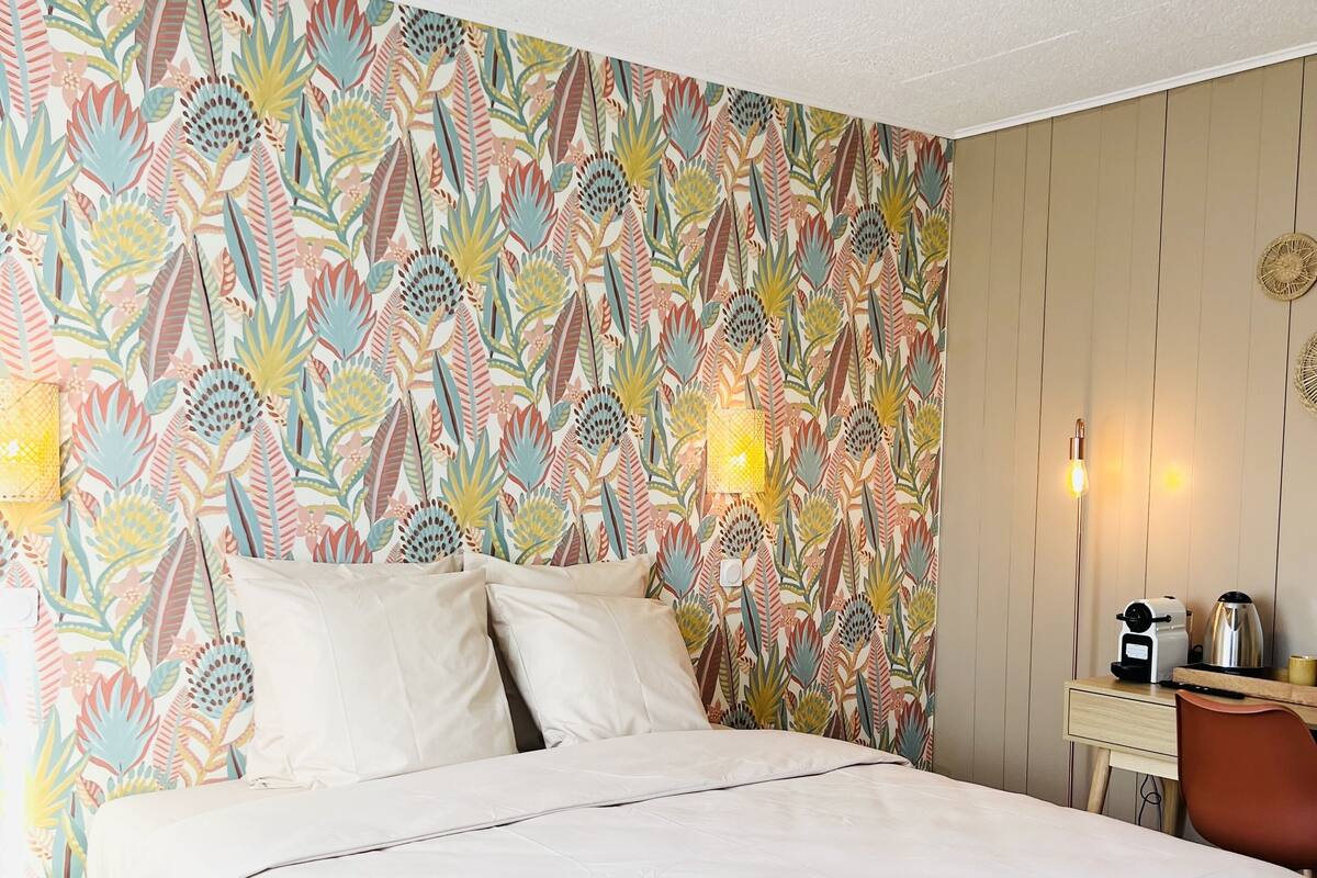 Un lit bien fait, recouvert d’une couvre-lit blanche, un mur orné d’un motif de feuilles colorées, une petite table de chevet avec une lampe, et un bureau en bois sur lequel se trouvent une bouilloire et une machine à café.