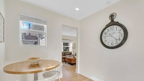 Un comedor con una mesa redonda de madera y sillas blancas, un reloj de pared y una ventana con persianas.