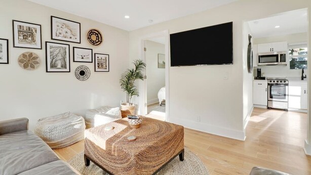 A modern living room with a large flat-screen TV, a coffee table, and a sofa. There is a kitchen area visible in the background. - Cottage on Croton Way # 2 (West Palm Beach)