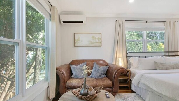 Un dormitorio con una cama, un sofá de cuero marrón, una mesa de centro con un florero y un cuadro en la pared.