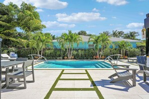 Una piscina con sillones de descanso y una mesa, rodeada de palmeras y un edificio al fondo.